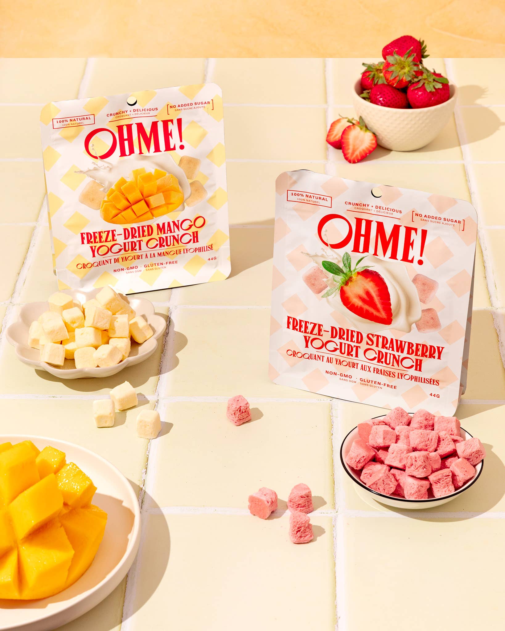 Two packages of OHME freeze-dried fruit snacks on a tiled surface with fruit and bowls.