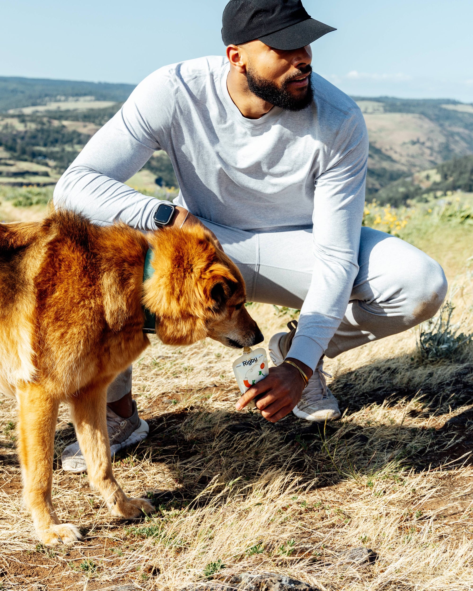 Man in a white shirt and black cap interacting with a brown dog who eating Rigby dog pouch treat on a hillside.
