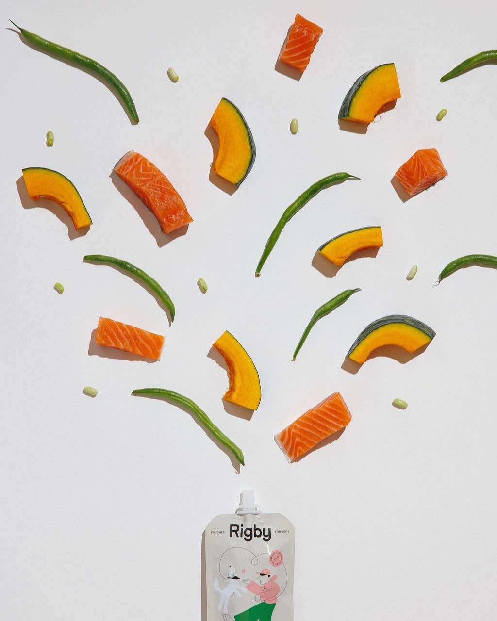 Fruit and vegetable slices arranged in a pattern with a 'Rigby' dog pouch treat product in the foreground.